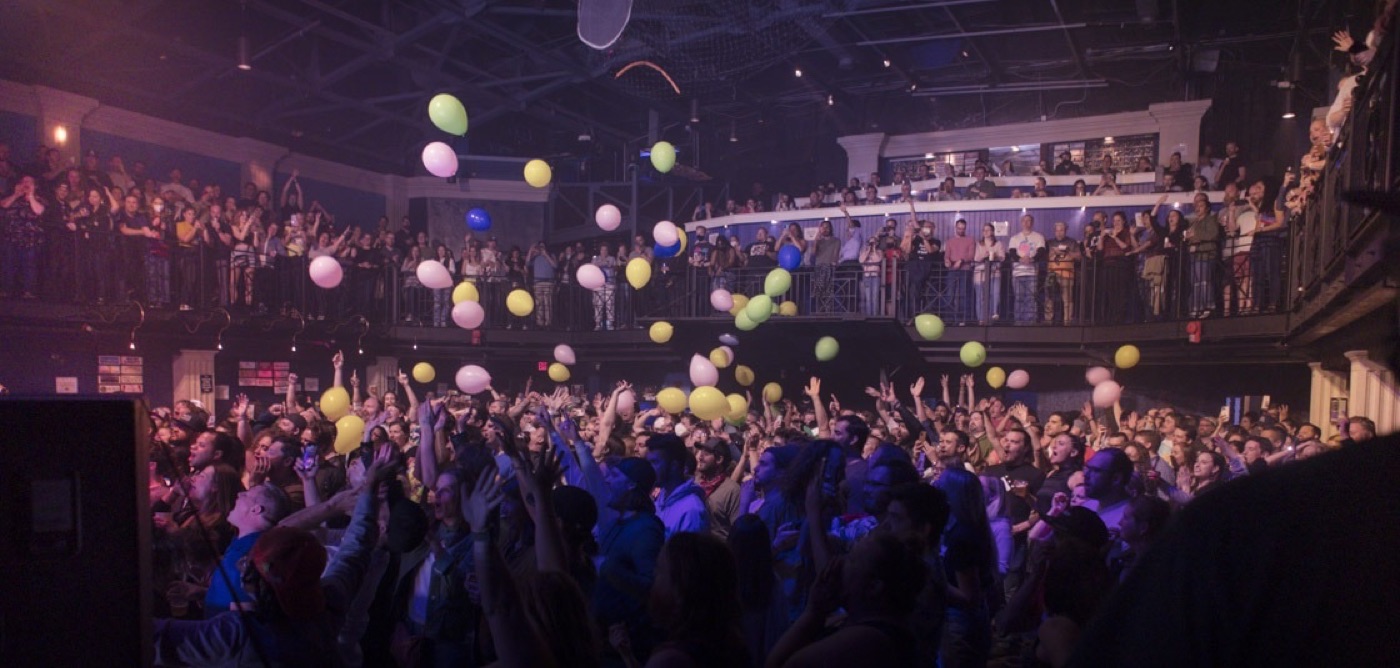 A packed club floor and balcony throw up their hands as dozens of pastel balloons fall over the crowd.