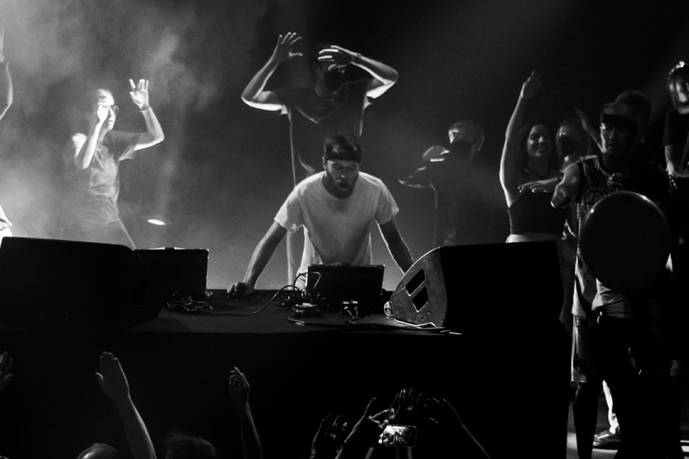 Black and white view of a bearded DJ in a headband leaning over his laptop while dancers throw up their hands on stage behind him.