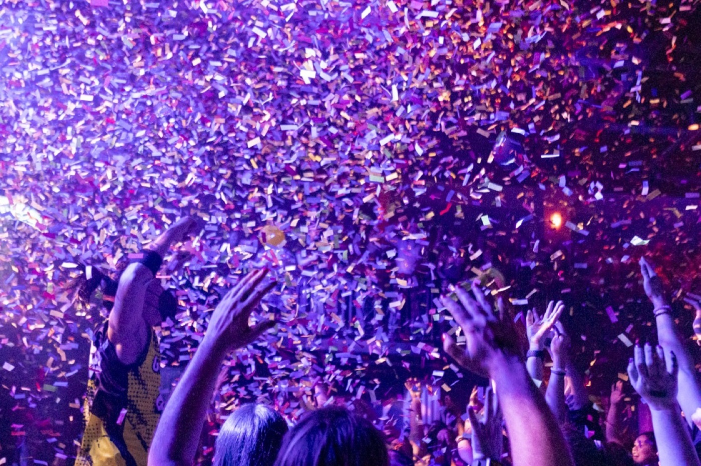 A storm of purple and gold confetti falls over raised hands at a concert.