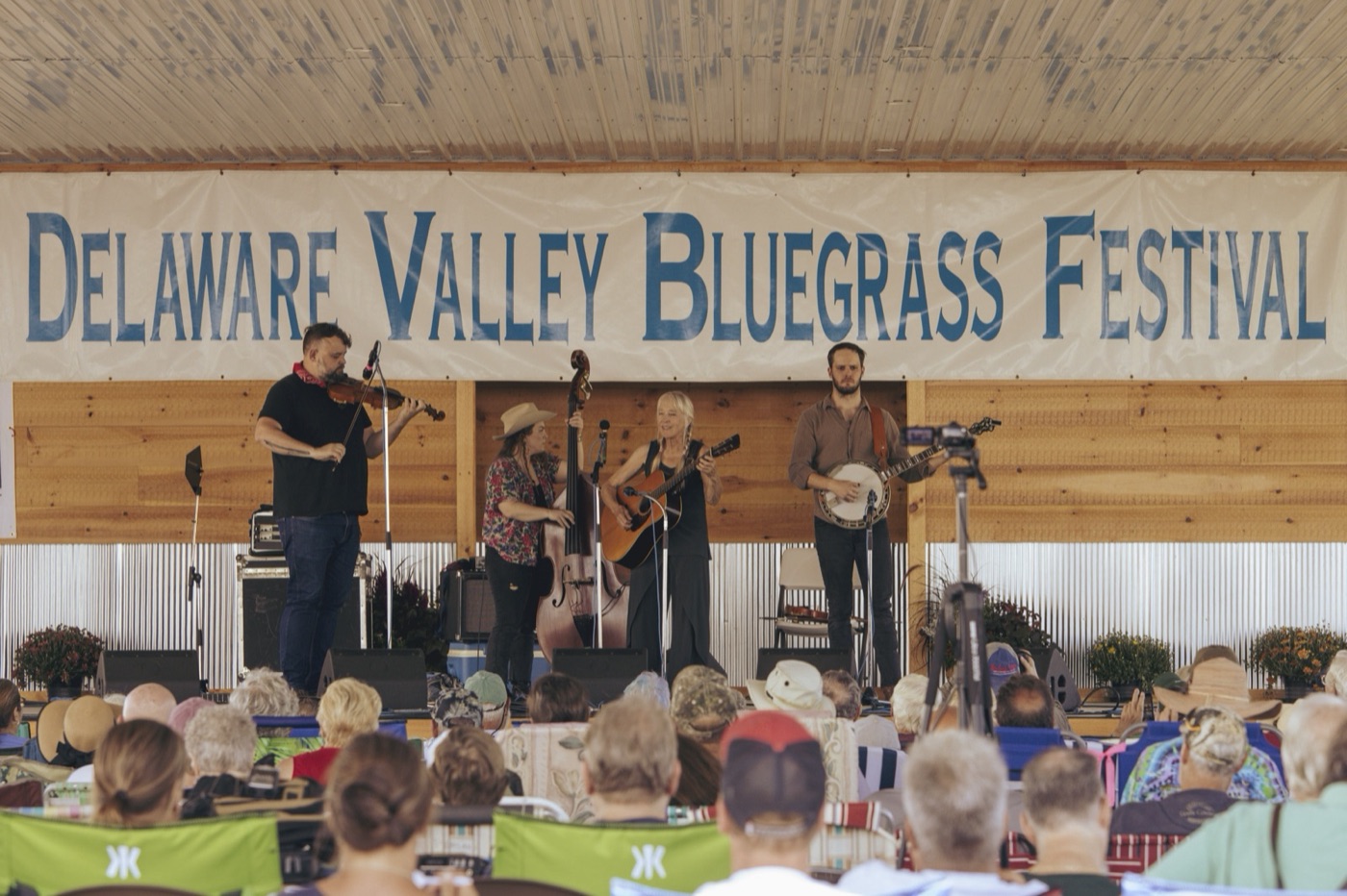 A four-piece bluegrass band with fiddle, upright bass, guitar and banjo plays under a Delaware Valley Bluegrass Festival banner to a seated crowd.