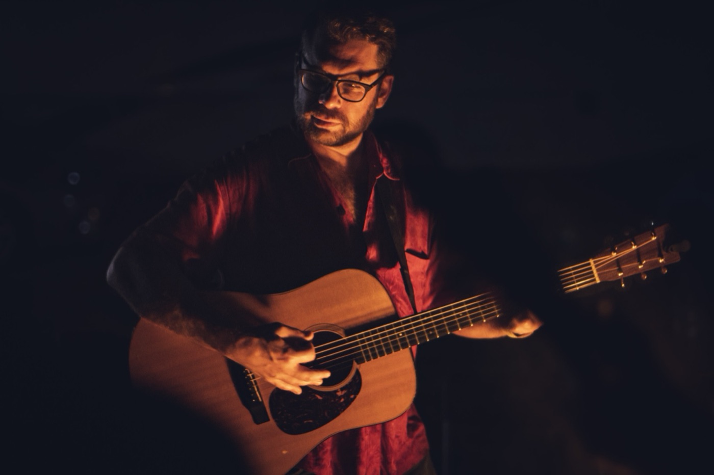 A bearded guitarist in glasses and a red shirt plays an acoustic guitar, lit warm against the dark at a night campground jam.