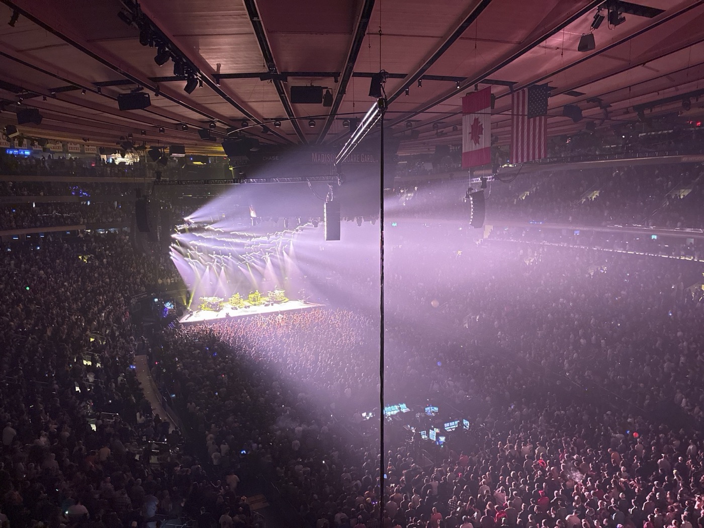 Pink and white light beams fan out from a far stage over a packed arena floor, with Canadian and American flags hanging from the roof.