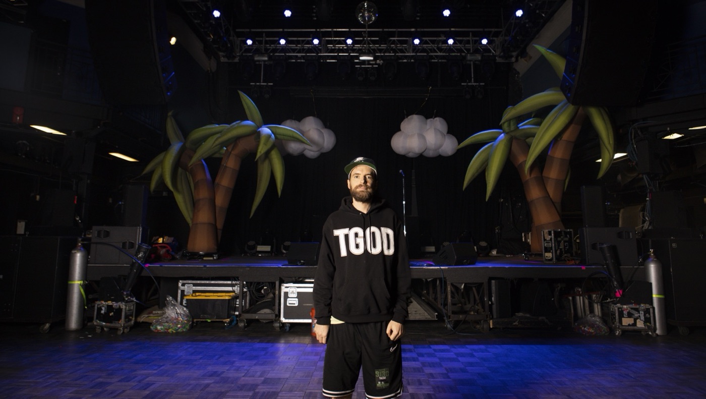 Girl Talk, in a black hoodie, stands alone on the empty stage before the show, between two inflatable palm trees.