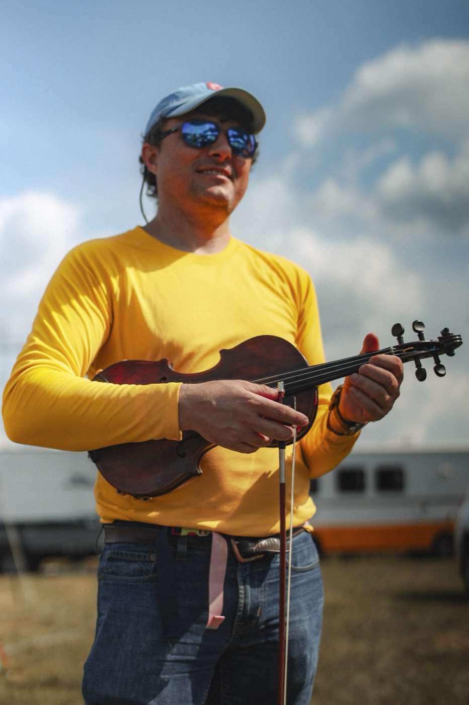 A fiddler in a yellow shirt and mirrored sunglasses holds his fiddle and bow under a summer sky.