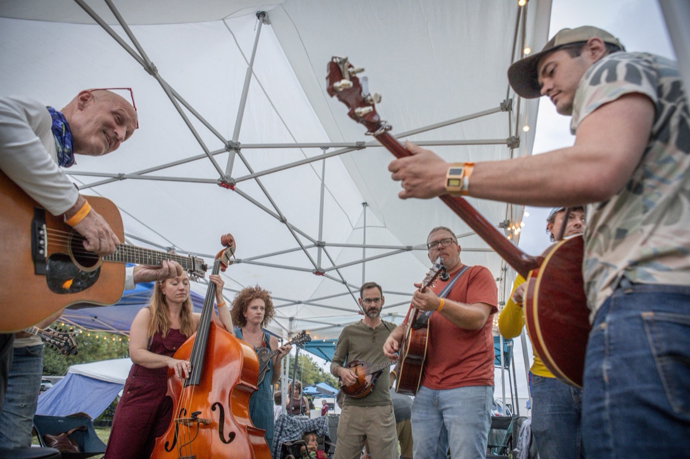 Bluegrass pickers with guitars, a mandolin and an upright bass jam in a circle under a white festival tent.