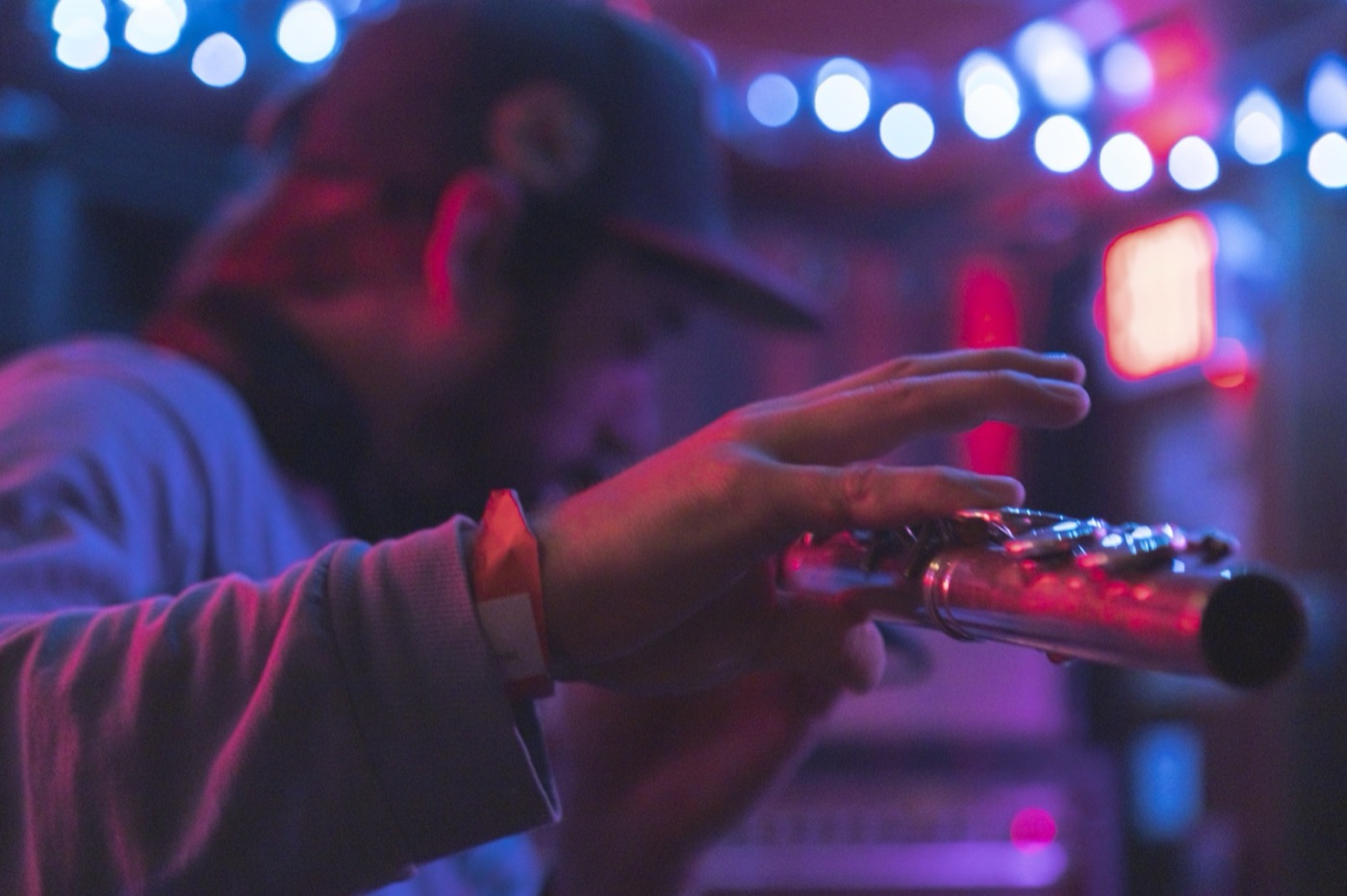 A musician holds a flute sideways, his hand sharp against blurred blue and red club lights.
