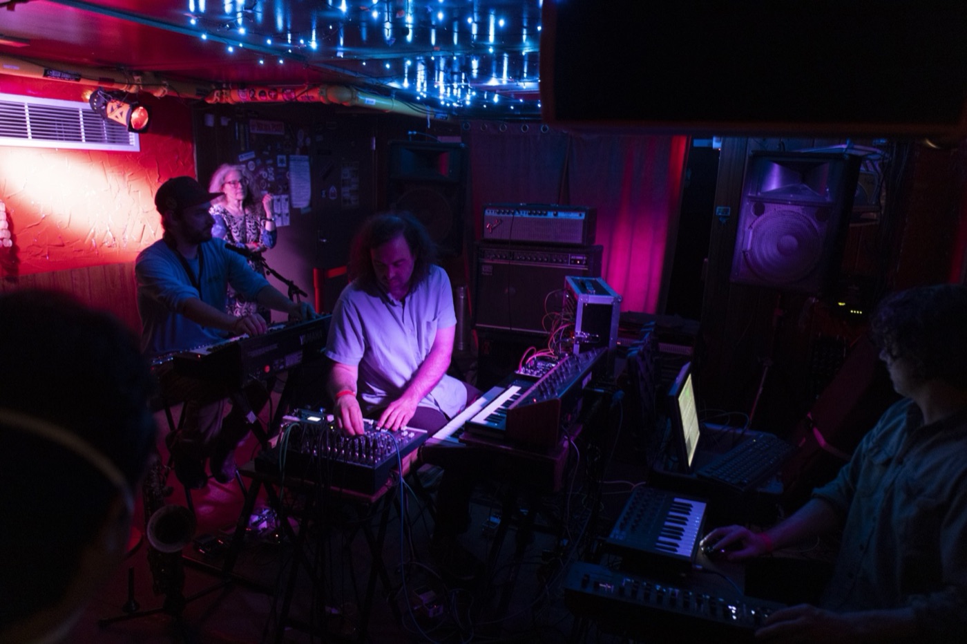 Three musicians play banks of synthesizers and keyboards in a small red club under strings of blue lights.
