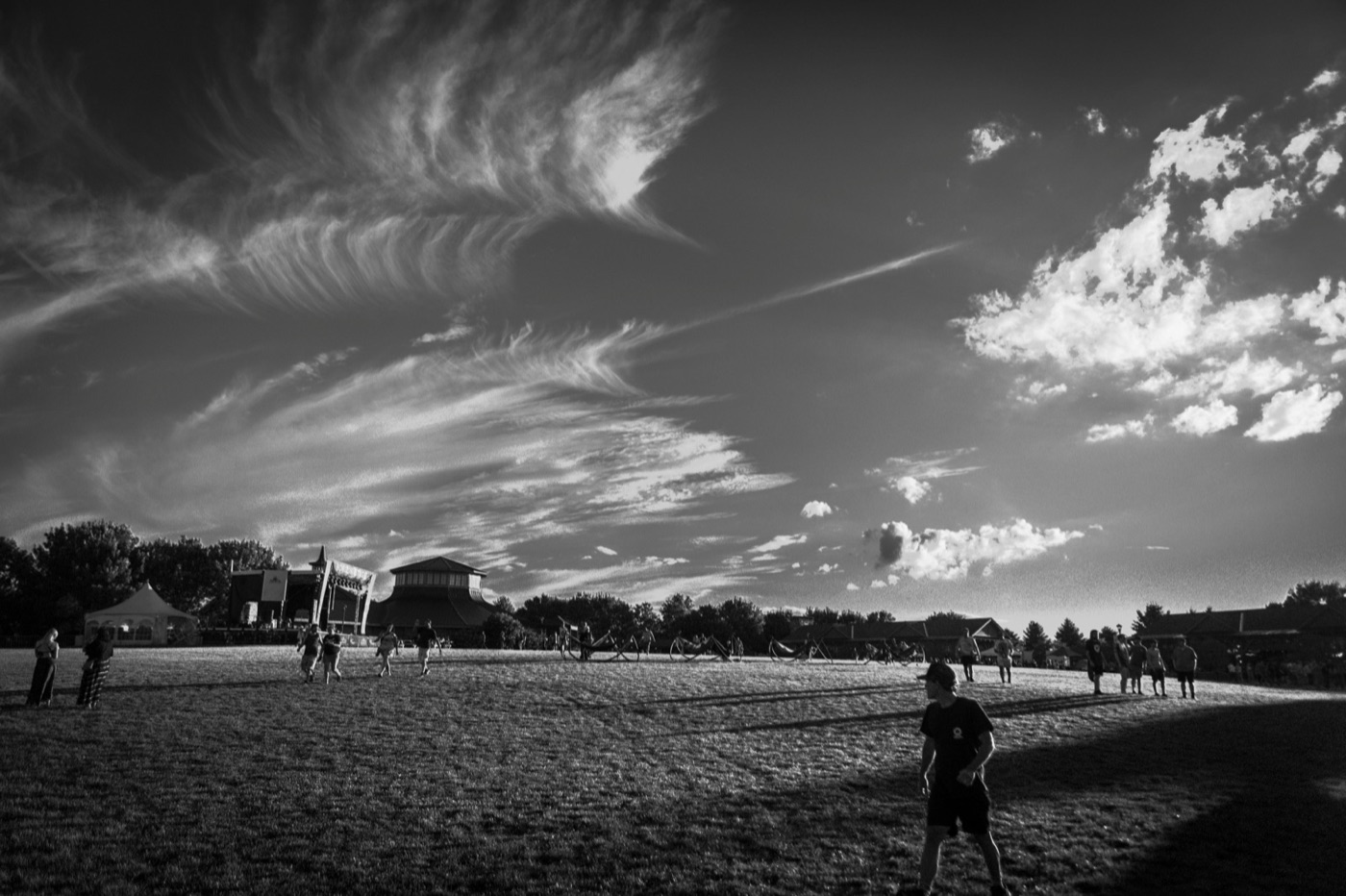 Black and white view of wispy clouds sweeping over a festival field with the concert pavilion in the distance.