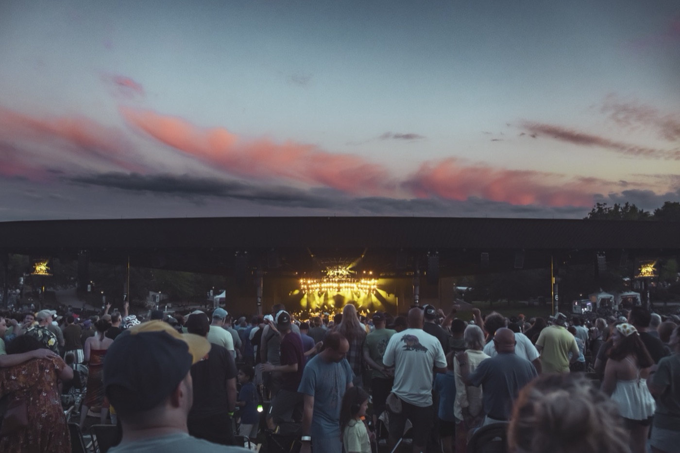 A packed lawn crowd faces a stage glowing gold under a pavilion roof while pink clouds streak the dusk sky.
