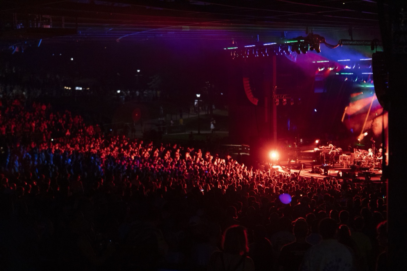 Red stage light washes over a packed crowd under a pavilion roof as the band plays at right.