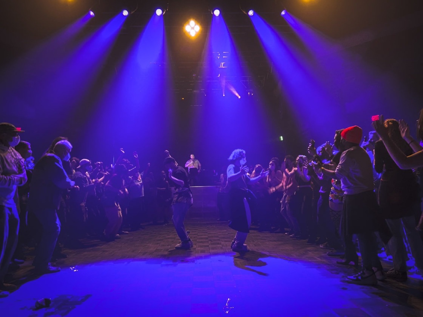 Blue light beams fan down over a ring of dancers on an open floor, with the performer at his table beyond them.
