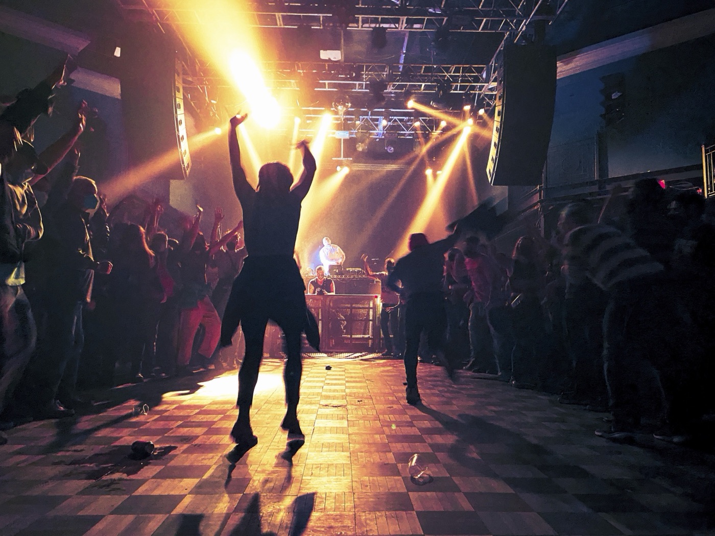 A dancer throws both arms up in silhouette against golden stage beams on a checkerboard dance floor, the performer lit at his table beyond.