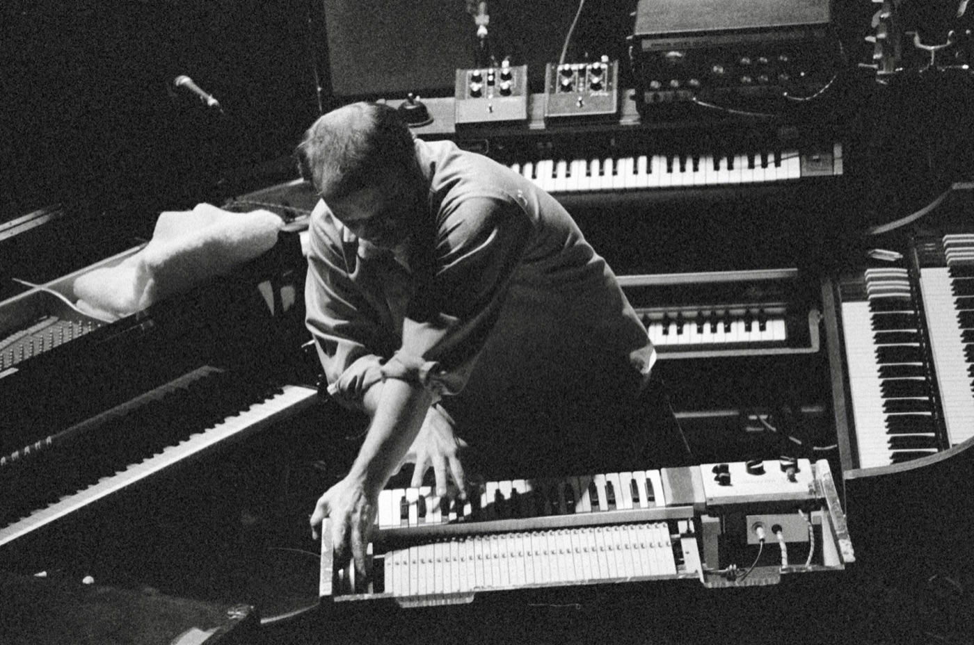 A keyboardist hunches low over a clavinet in the middle of a ring of keyboards, shot from above in black and white.