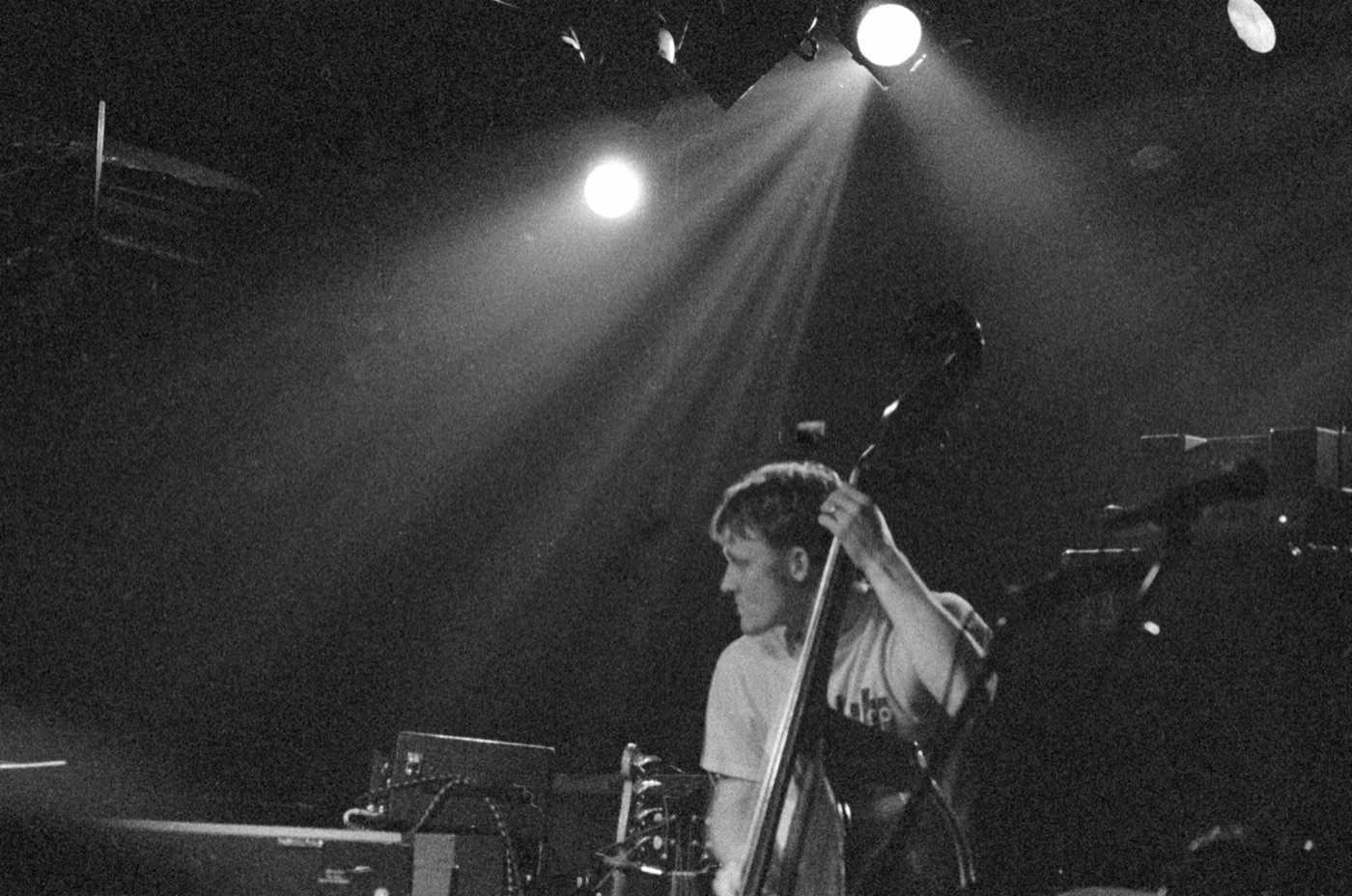 A bassist plays upright bass under spotlight beams cutting through a dark, smoky stage in black and white.