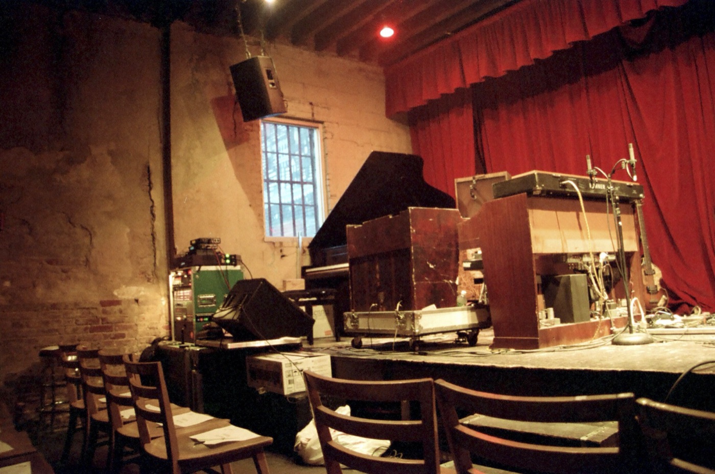 The inside of a small brick club, rows of wooden chairs facing a stage crowded with a piano, organ and gear under a red curtain.