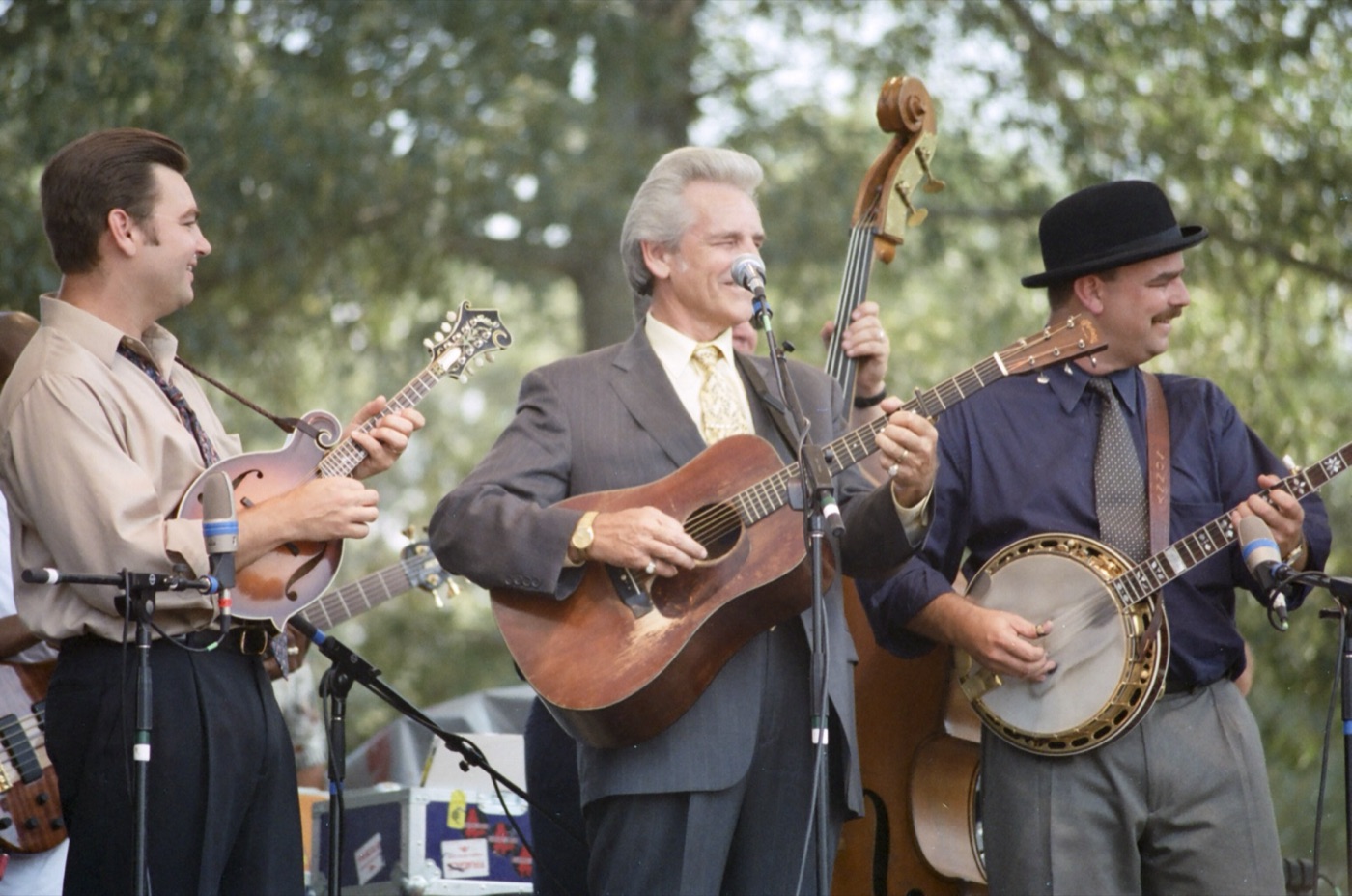 Three suited bluegrass players on mandolin, acoustic guitar and banjo grin at one another on an outdoor stage under trees.
