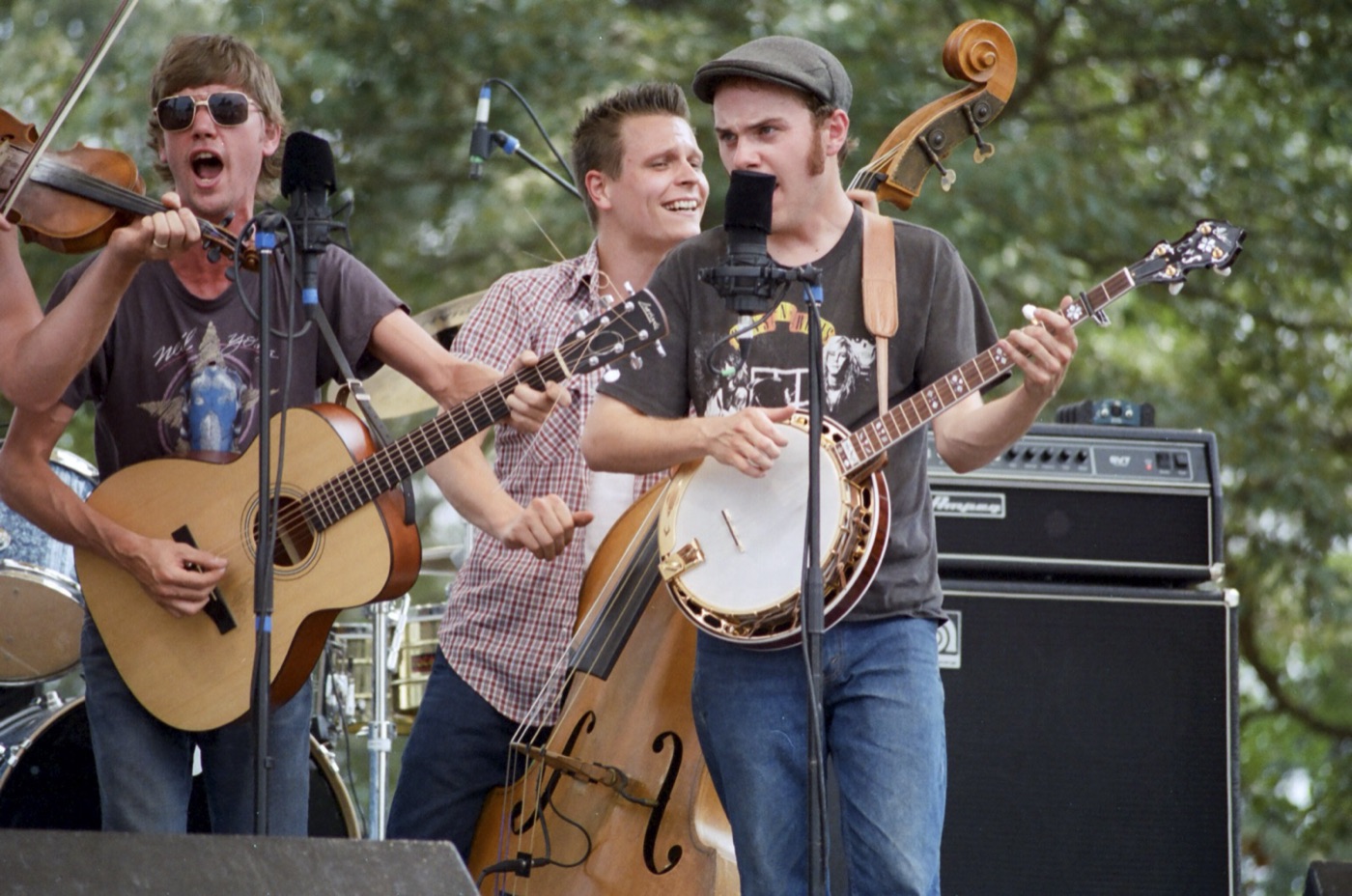 A fiddler, a guitarist and a banjo player sing and grin at their microphones, with an upright bass behind them.