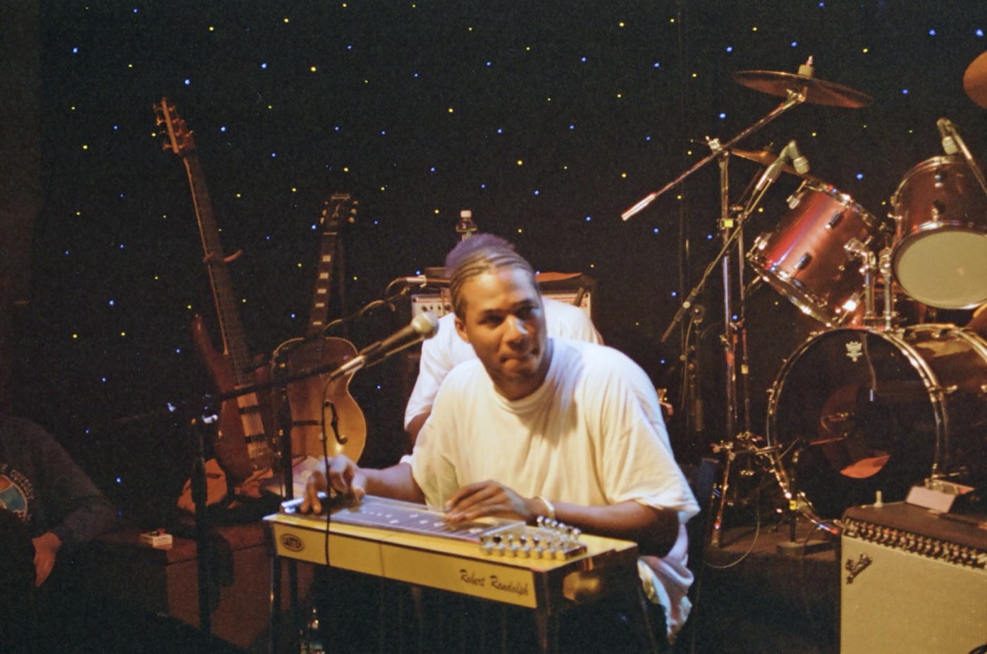 A pedal steel player in a white T-shirt looks up from his instrument in front of a drum kit and a starry backdrop.