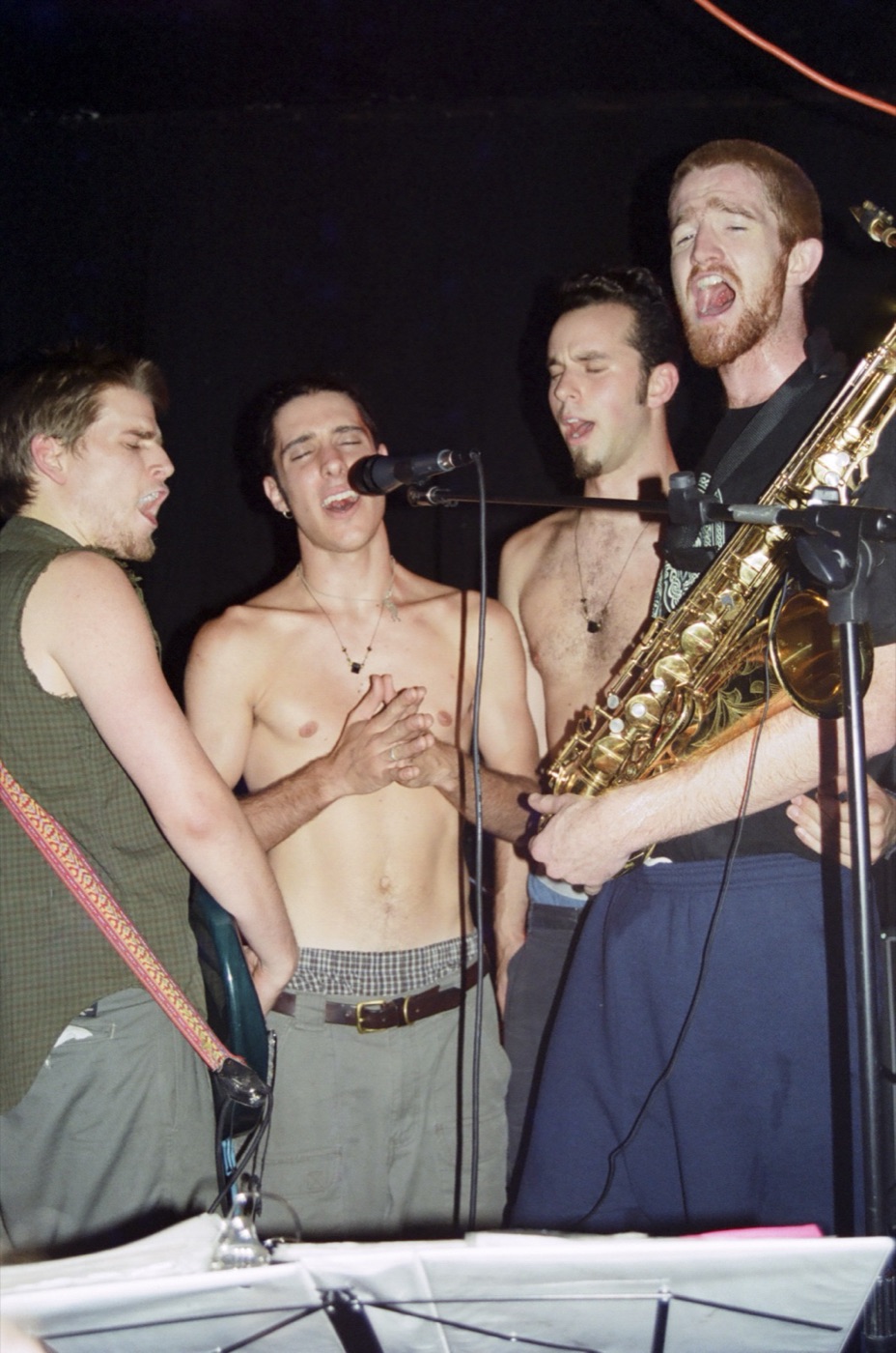 Four band members crowd one microphone and sing with eyes shut, a saxophone in front.