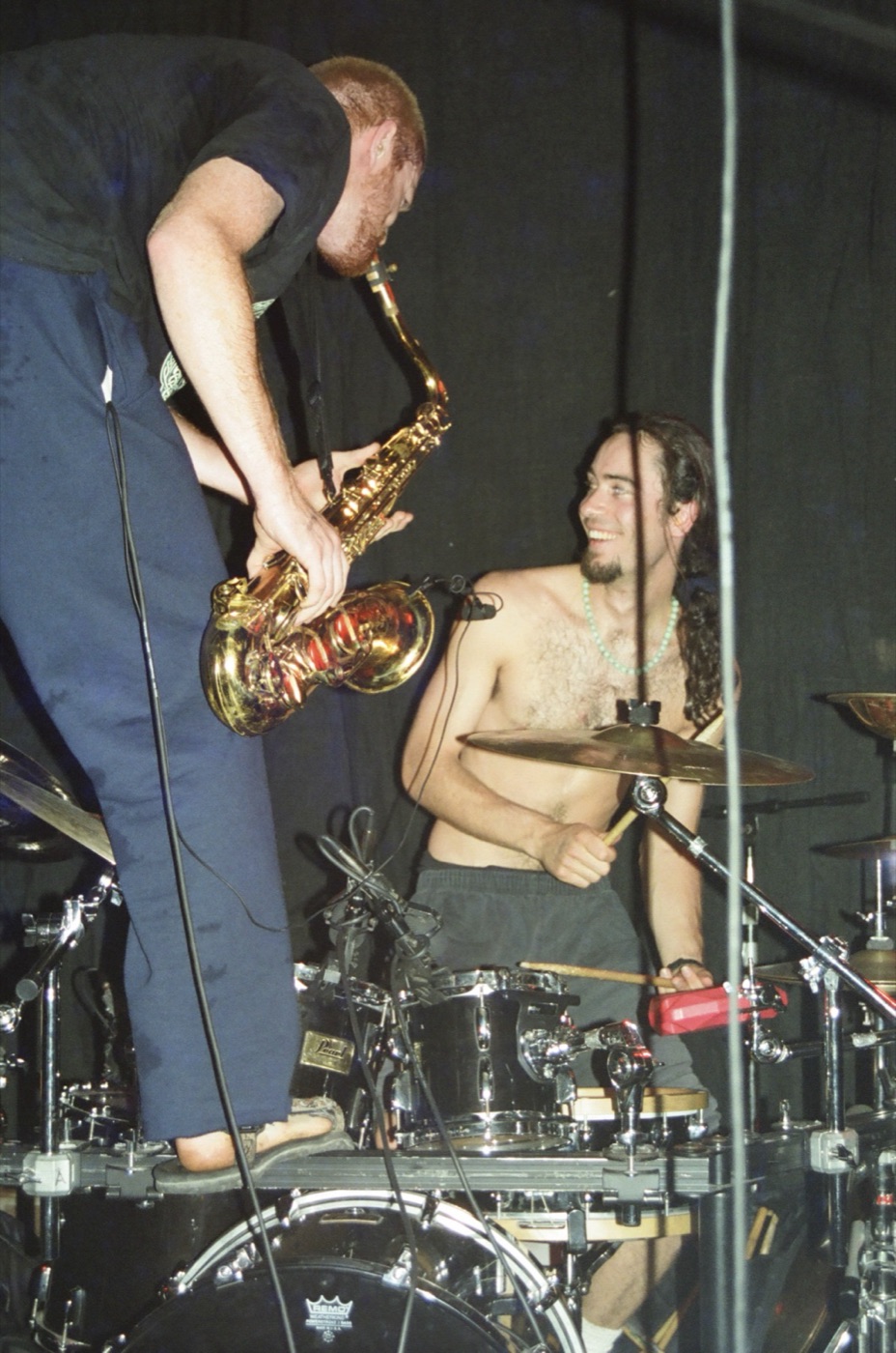 A saxophonist stands on the bass drum and leans over a grinning shirtless drummer mid-song.