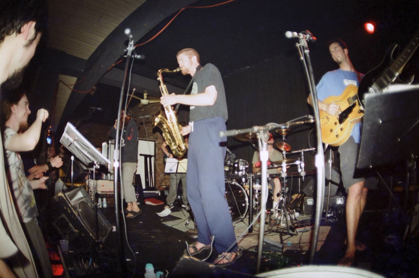 A wide fisheye view of a small club stage with a saxophonist, guitarist and drummer as fans cheer at the edge.