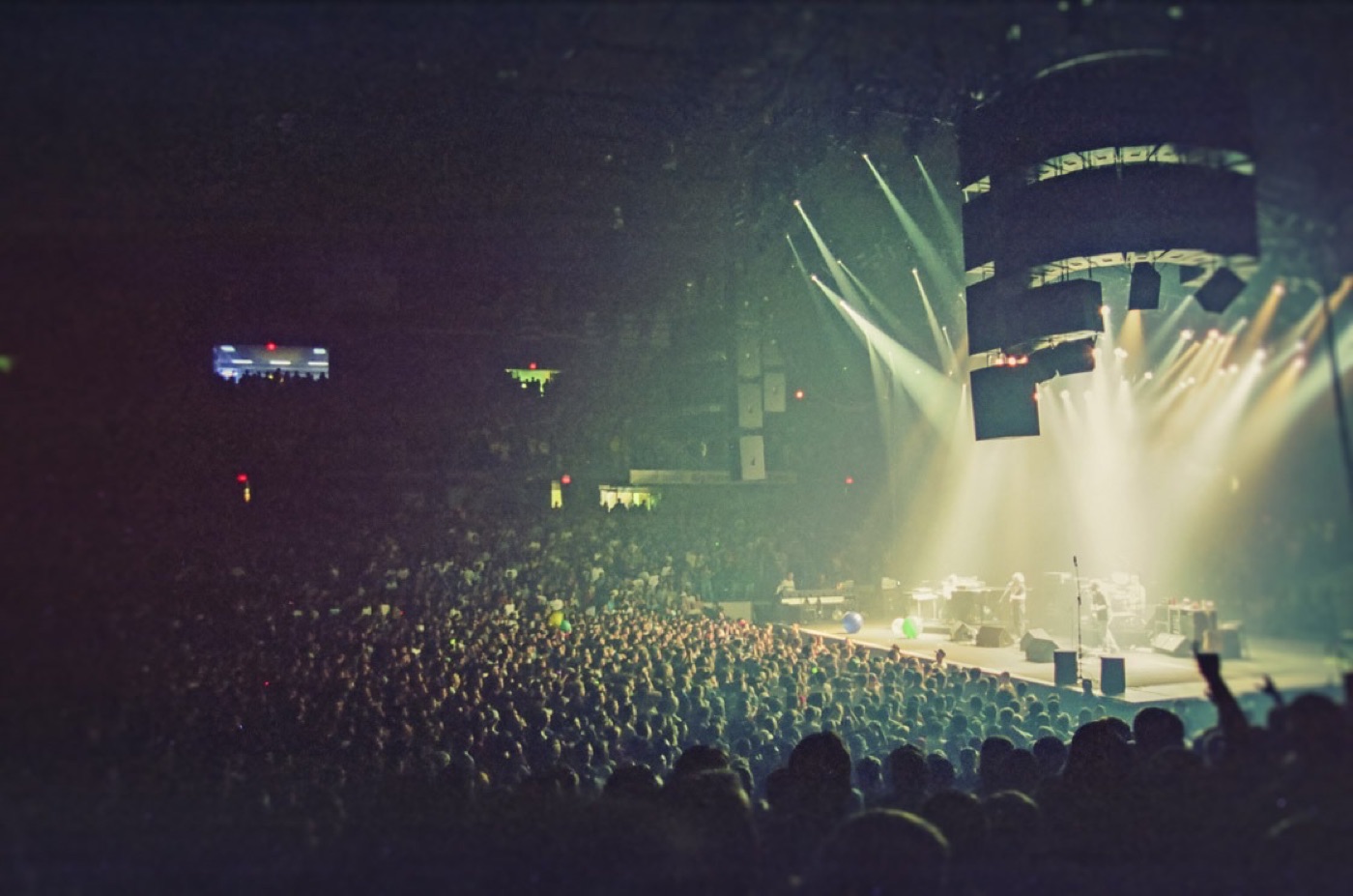 A packed arena crowd faces a distant stage washed in pale green light beneath a hanging speaker array.