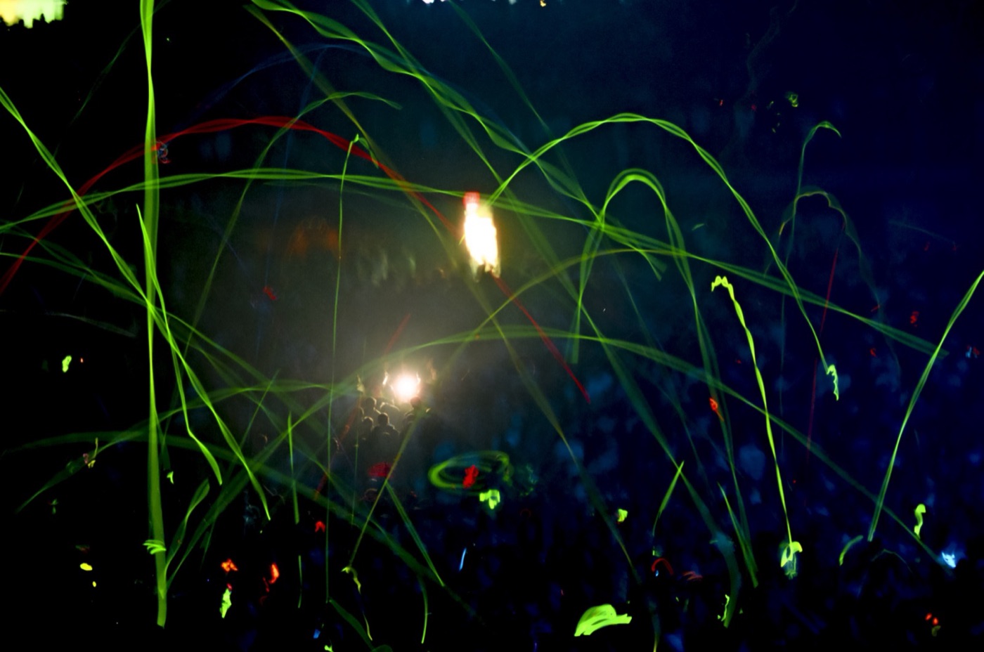 Glow sticks streak in green arcs over a dark arena crowd as a spotlight burns through the haze.