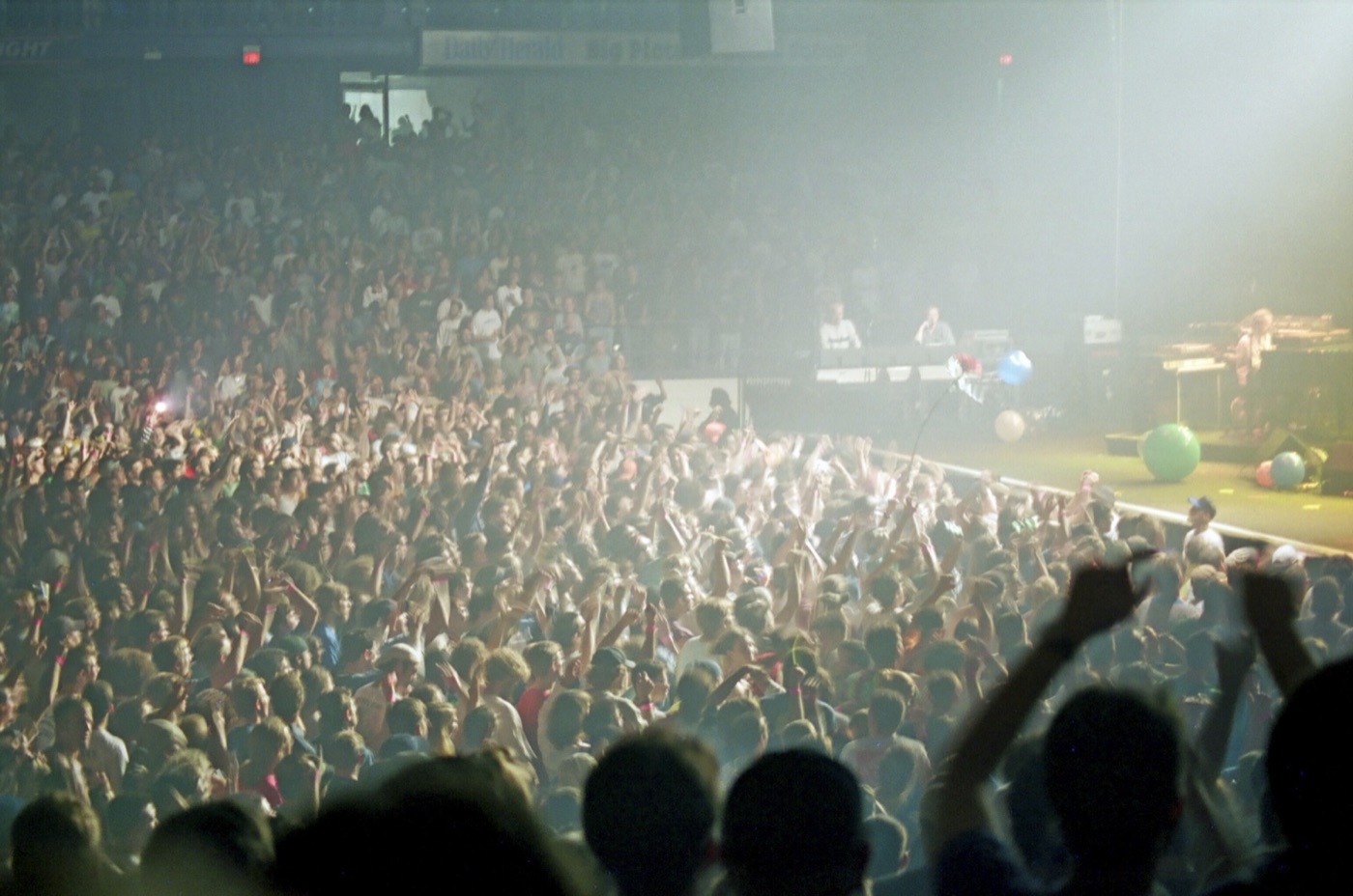 A hazy arena floor full of fans with arms raised toward a stage where beach balls roll by the monitors.
