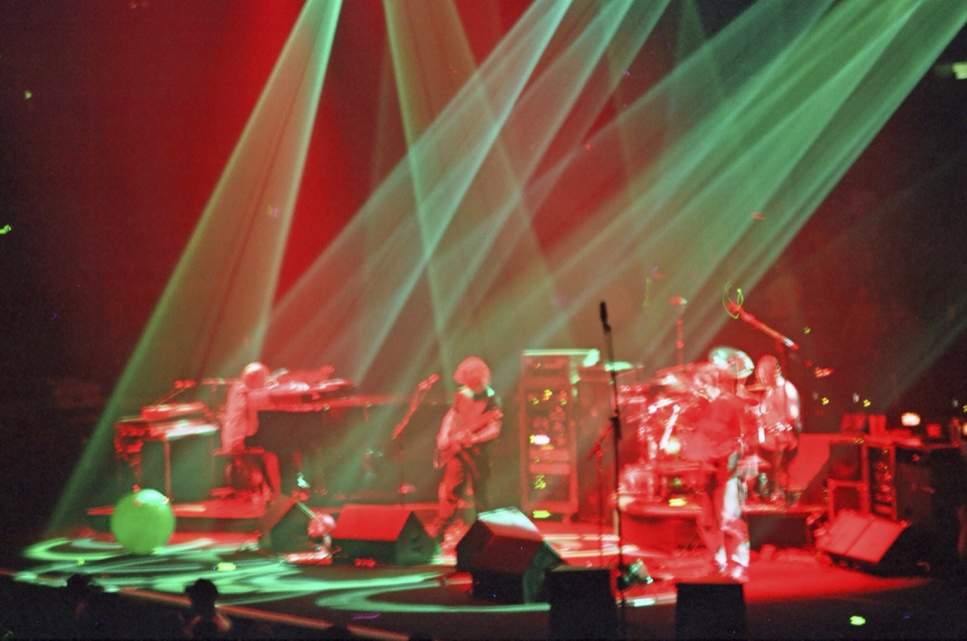 A band plays keys, guitar and drums on a stage flooded red and crossed by green light beams.