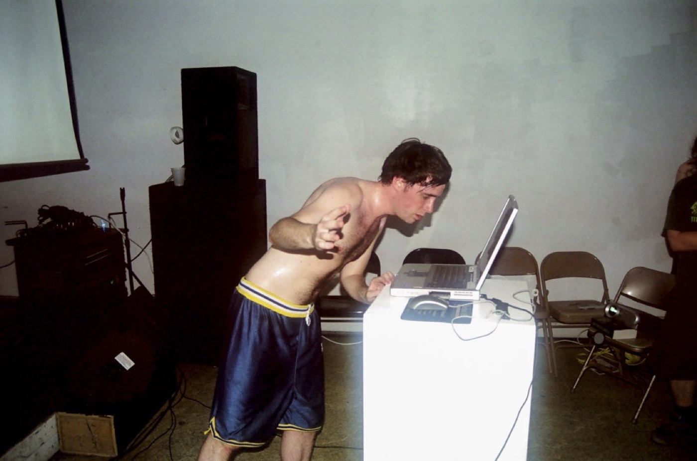 A shirtless, sweaty performer in basketball shorts leans into a laptop on a white plinth, with a speaker stack and a projection screen behind him.