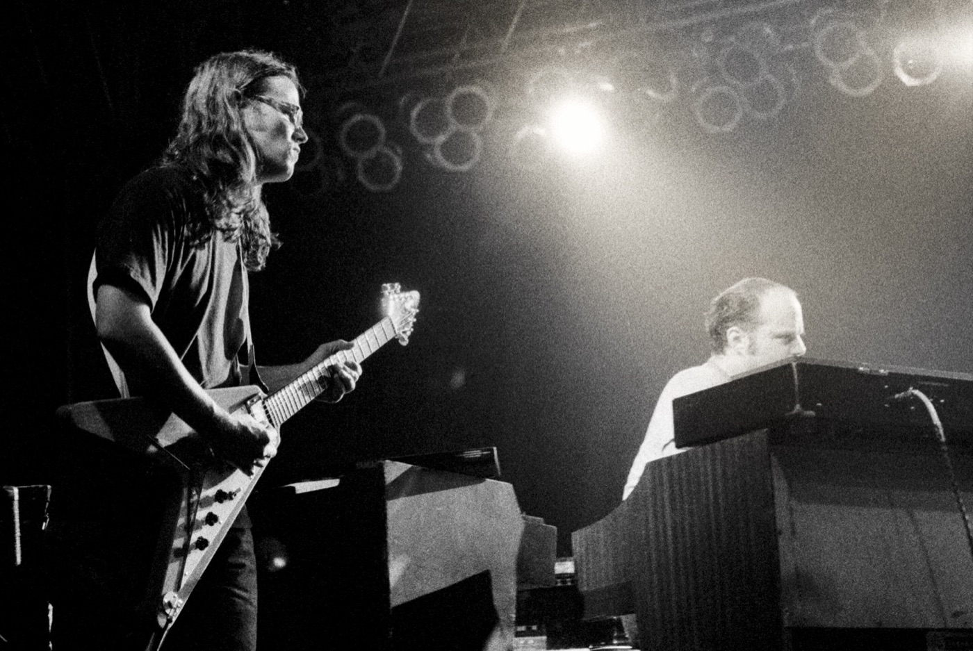 In black and white, a long-haired guitarist with a Flying V plays beside the organist under a hanging stage light.