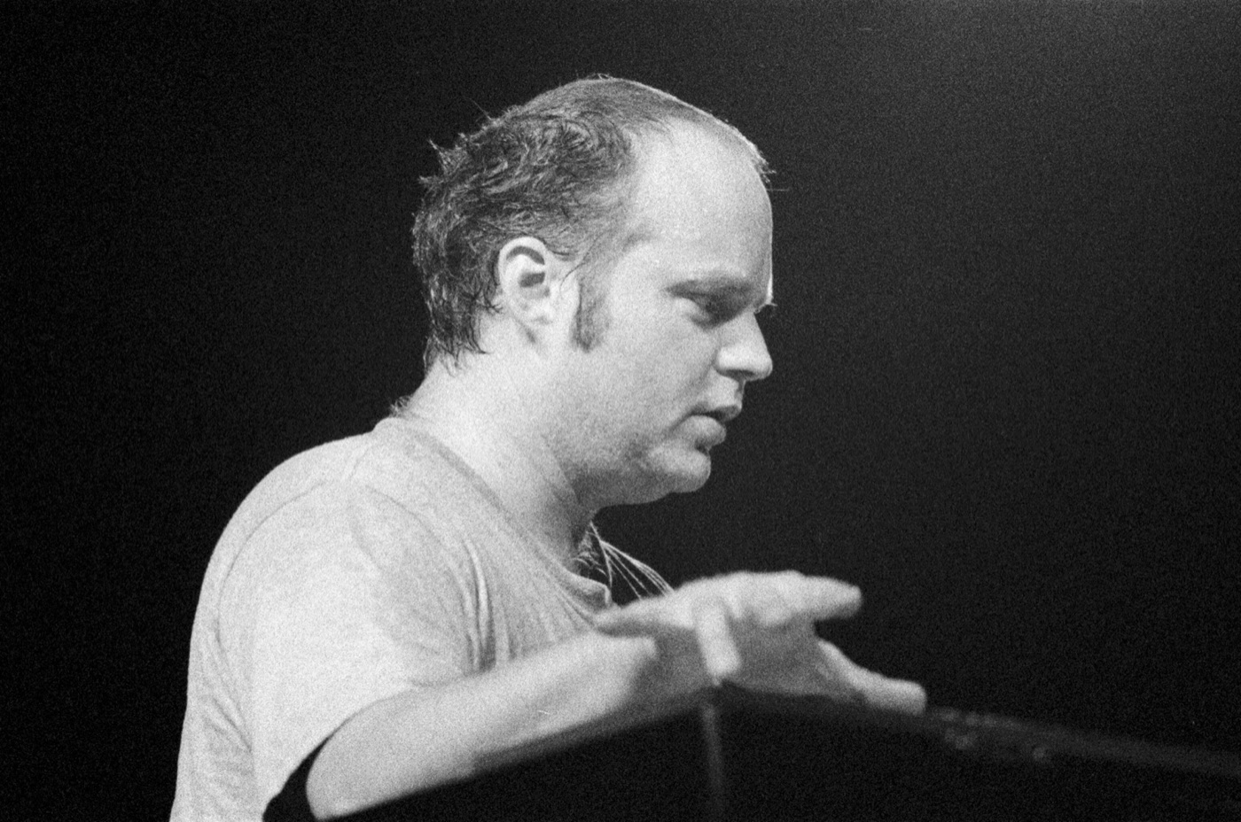 In black and white, the organist plays in profile with his hands raised over the keys against a black background.