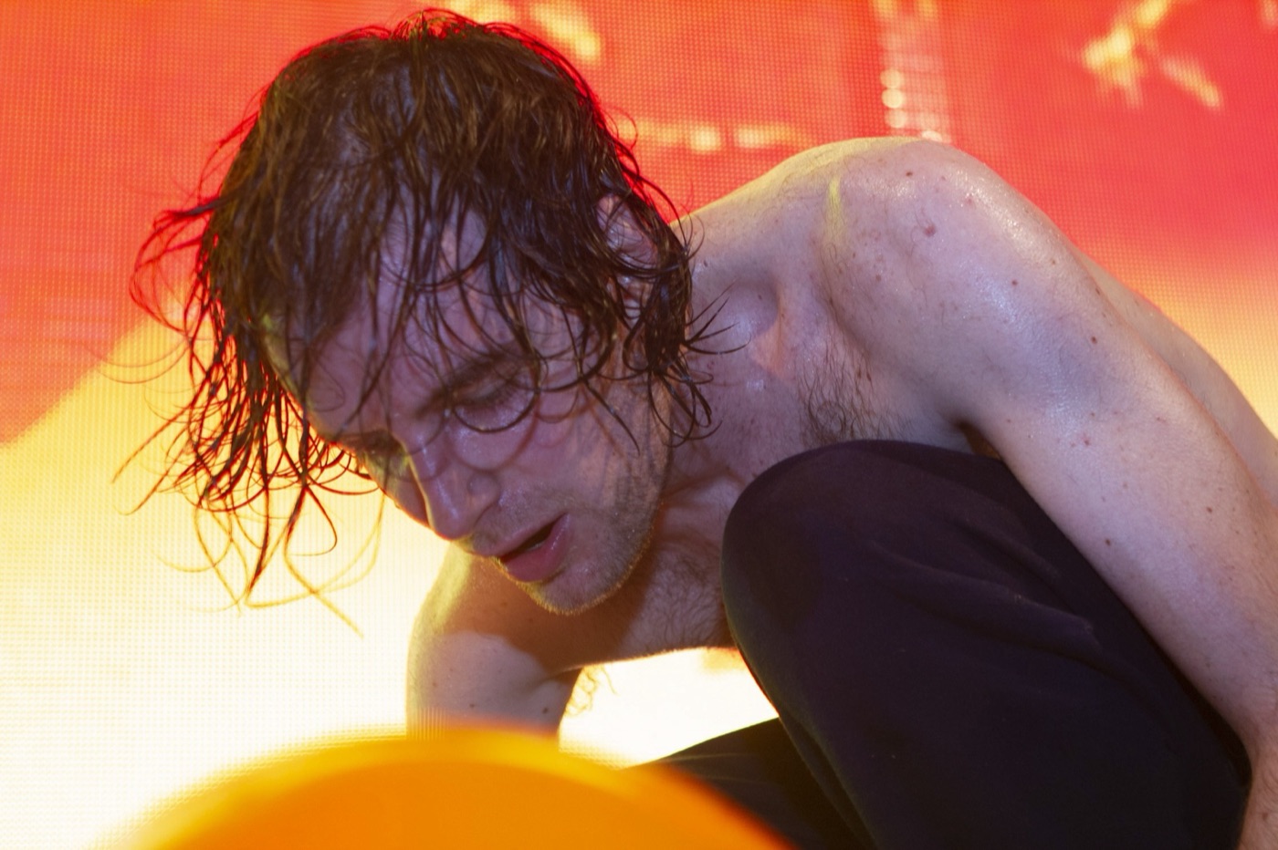 Girl Talk crouches on stage with dripping wet hair against a blazing orange screen, a balloon in the foreground.