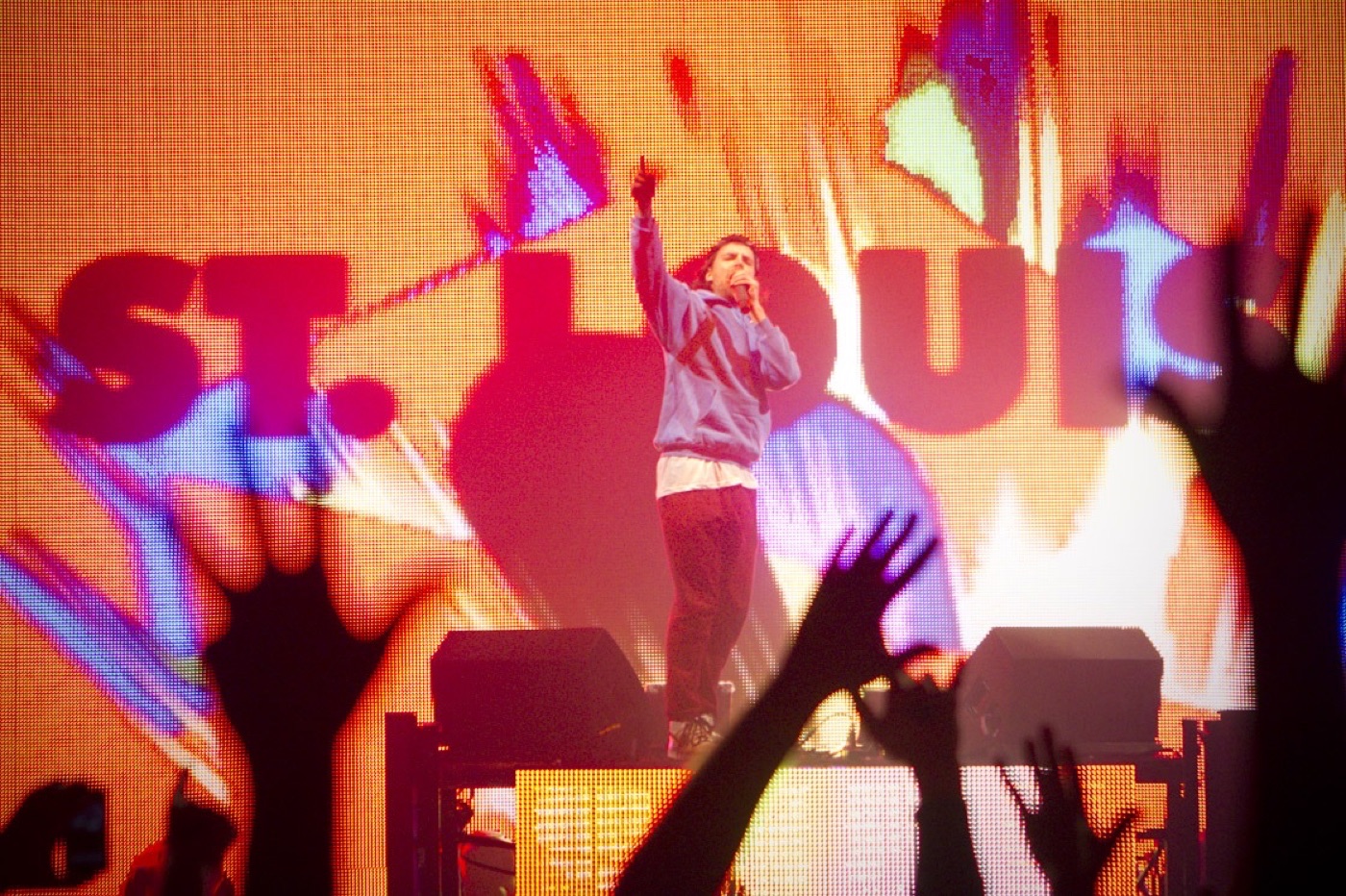 Girl Talk points skyward with a microphone in front of a giant orange and pink ST. LOUIS video wall as fans raise their hands.