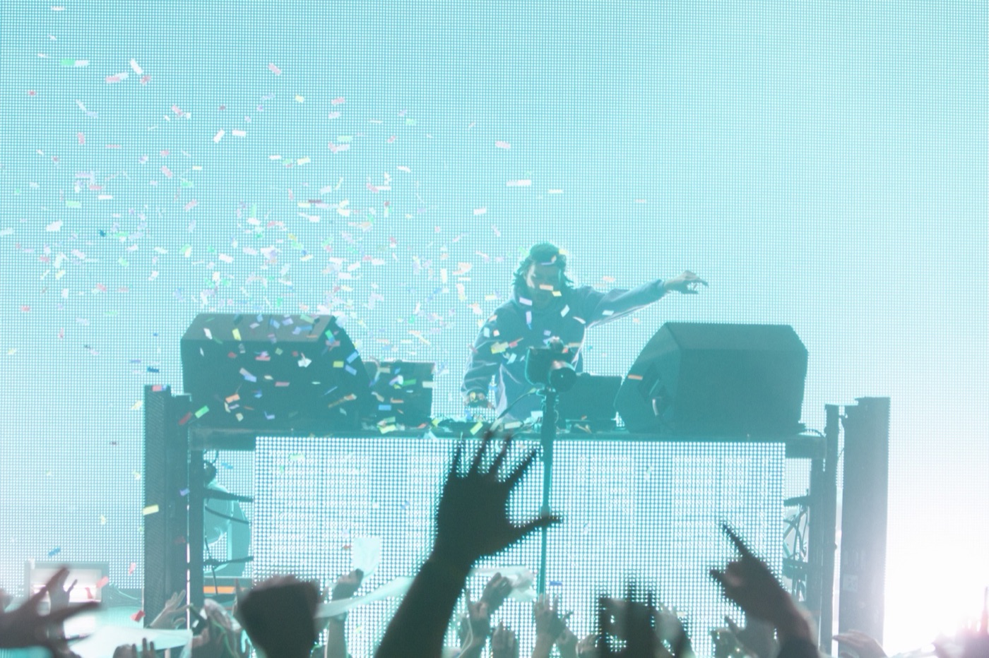 Confetti rains against a cyan screen as the DJ flings an arm out over the booth and the crowd's hands reach up.